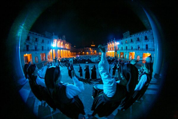 Vista gran angular de músicos tradicionales y asistentes en una plaza histórica iluminada durante un evento nocturno.